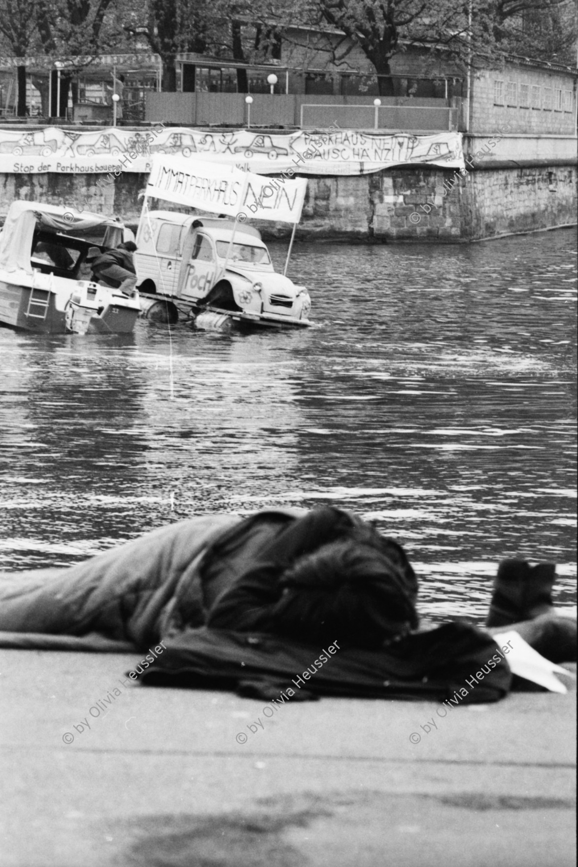 Image of sheet 19800080 photo 7: Mann schlaeft an der Rivi während Demonstration mit Citroen auf Floss der POCH gegen das Limmatparking, Zuerich 1980. 

Protest Limmat