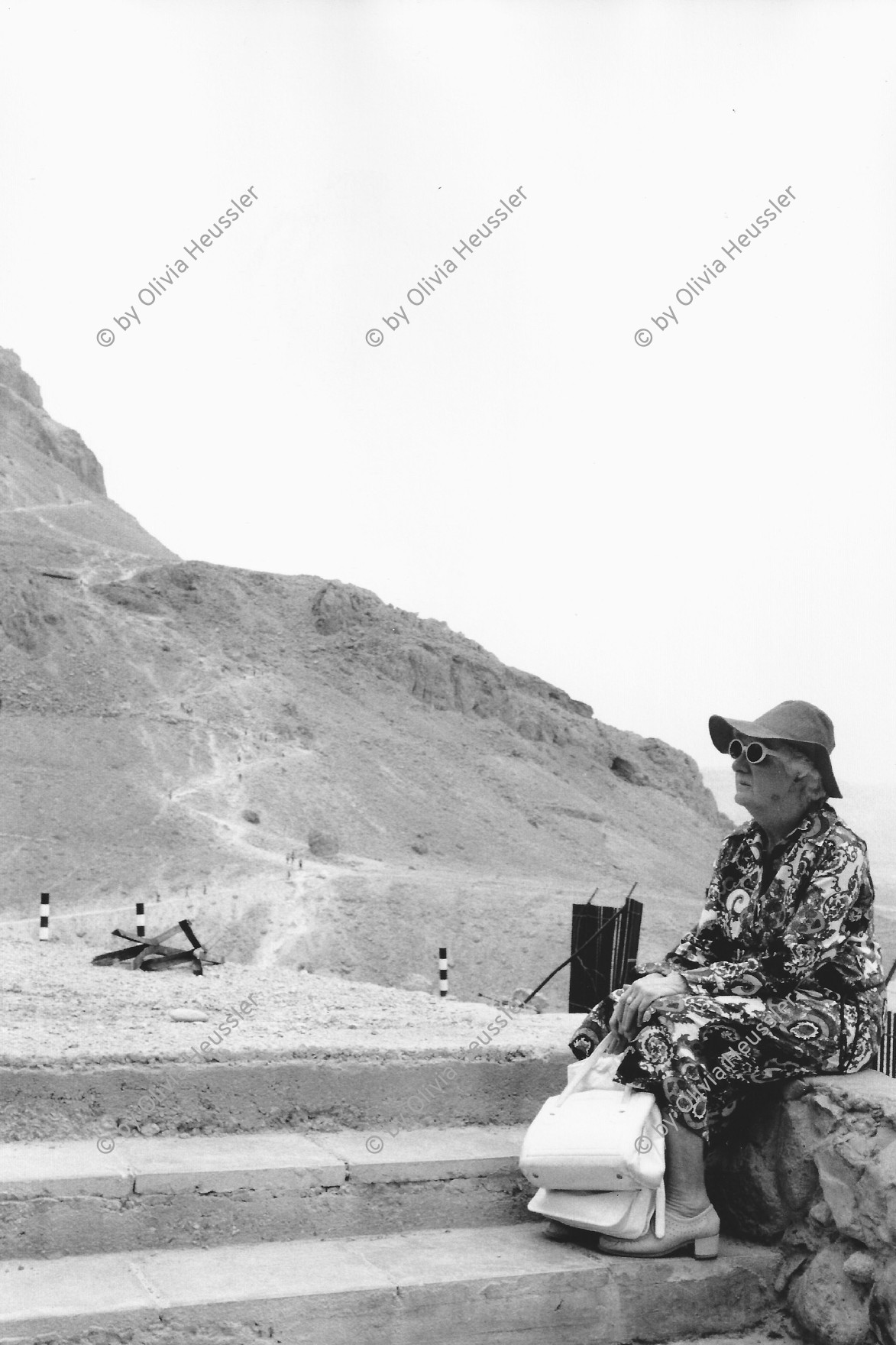 Image of sheet 19800120 photo 7: Eine Seniorin und Touristin ruht sich aus. Massada Israel. Palestine 06.80 1980 √ 
An elderly tourist takes a rest. resting turist sunglasses and sun hat bags sitting lady 
Aus: Out of Jerusalem / Jenseits von Jerusalem page 87

Masada (Hebrew מצדה, pronounced About this sound Metzada (help·info), from מצודה, metzuda, "fortress") is an ancient fortification in the Southern District of Israel, on top of an isolated rock plateau (akin to a mesa) on the eastern edge of the Judaean Desert, overlooking the Dead Sea. Herod the Great built palaces for himself on the mountain and fortified Masada between 37 and 31 BCE. The Siege of Masada by troops of the Roman Empire towards the end of the First Jewish–Roman War ended in the mass suicide of the 960 Jewish rebels holed up there. Masada is located 20 kilometres (12 mi) east of Ara