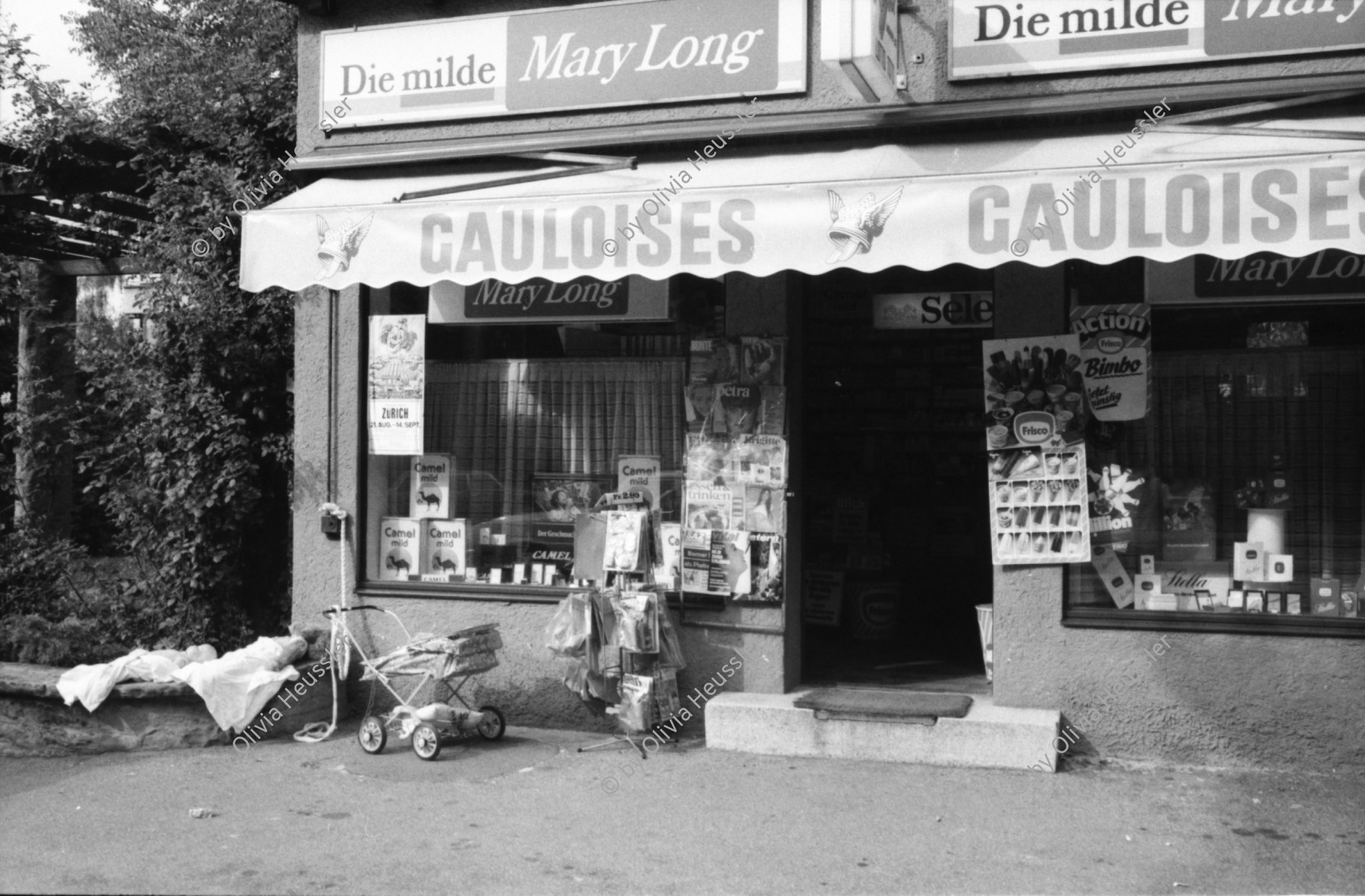 Image of sheet 19800400 photo 7: Kinderwagen  und Spielsachen vor einem Kiosk. Güterwagen überqueren die Strasse.