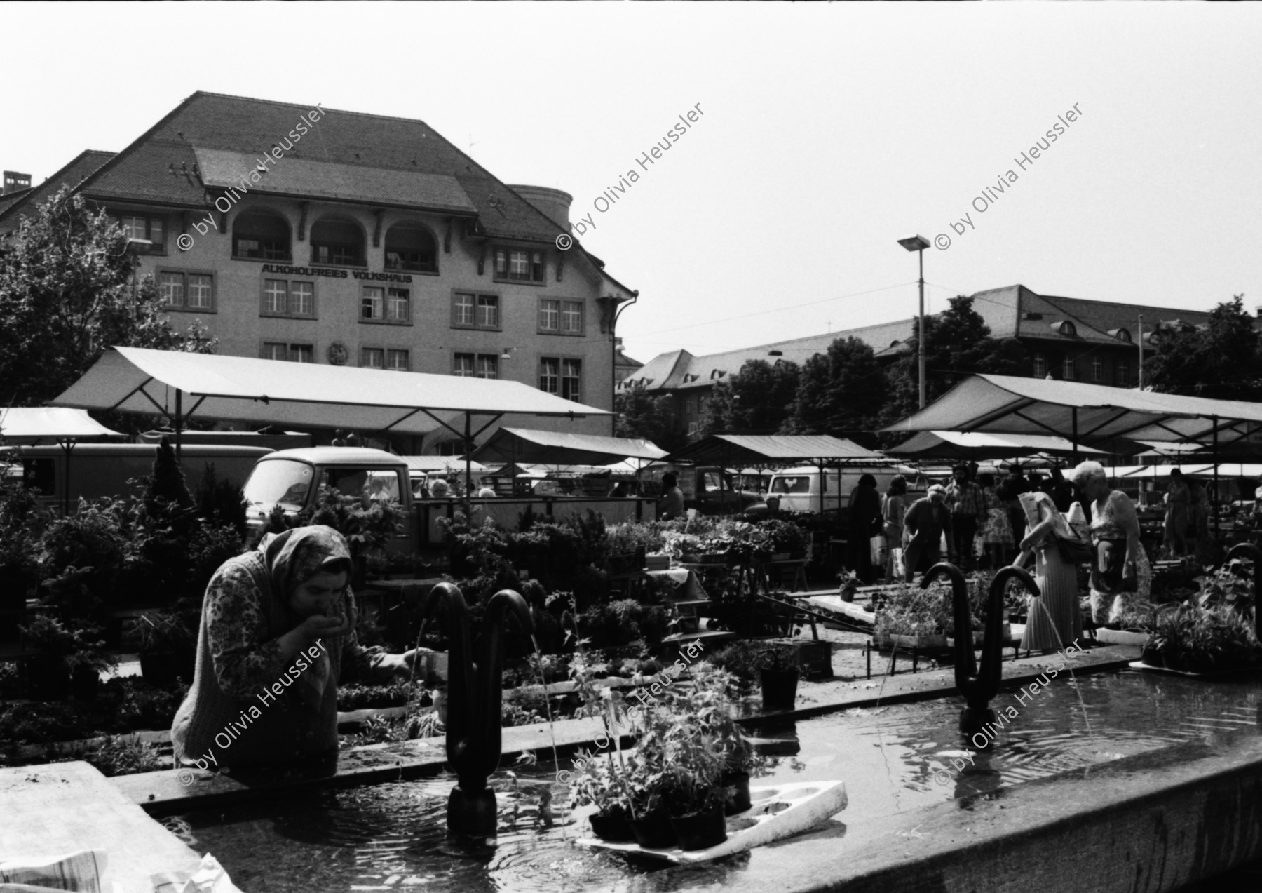 Image of sheet 19810210 photo 4: Frau trinkt am Brunnen Wasser. Marktstimmung auf dem Helvetiaplatz. Zürich vor dem Volkshaus. Kreis 4 Aussersihl Einkaufen Markt