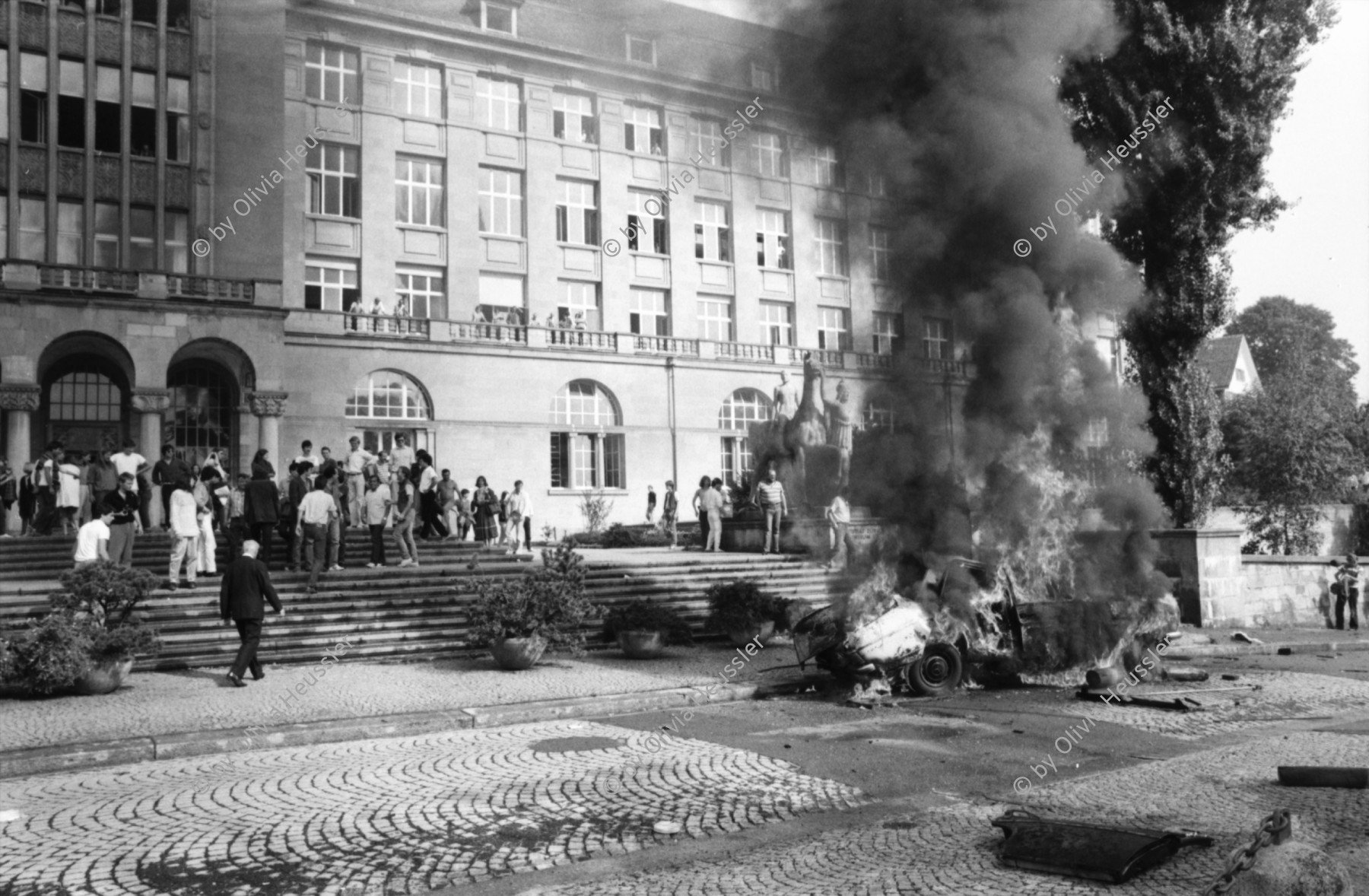 Image of sheet 19810450 photo 17: Drehaufnahmen zum Film Espion leve toi von Yves Poisset 
Männer befestigen Schweizerfähnchen auf Mercedes Benz
brennt noch Loch im Schlauch der Feuerwehr Rauchfahne
Zürich Künstlergasse 1981