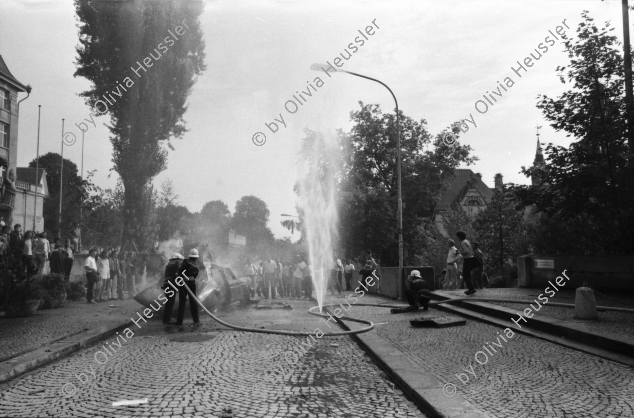 Image of sheet 19810450 photo 20: Drehaufnahmen zum Film Espion leve toi von Yves Poisset 
Männer befestigen Schweizerfähnchen auf Mercedes Benz Universität Zürich
brennt noch Loch im Schlauch der Feuerwehr Rauchfahne
Zürich Künstlergasse 1981