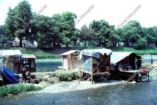 Image of sheet 19813002 photo 5: Chaotikon in der Sihl. Protest gegen Wohnungsnot Wohn Installation Zürich occupied house √
Barraken siedlung auf Pfosten, Küche Kreis fünf 1981 Wohnen housing banner river protest settlement Schweiz 
Heinz Nigg:
31. April 1981
Bewegte beginnen mit dem Bau von Neu-Chaotikon auf einer Insel in der Sihl in unmittelbarer Nähe des wiedereröffneten AJZ.
19. Mai 1981
Die Polizei räumt Neu-Chaotikon. Aus Protest gegen die Räumung beginnen Bewegte am Abend, den Belag des Car-Parkplatzes neben dem AJZ mit Pressluftbohrern aufzureissen. Die Polizei greift ein.