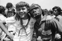 Image of sheet 19840290 photo 10: Geburtstag Sandinos. Des Generals des freien Menschen, wird gefeiert. «Sandino vive» steht an den Mauern. Kinder in Reihe. Gruppenbild. Bühne. FSLN Fahne. Mann Soldat mit Maske. Kommandanten mit Strohhüten. Fahnen. Mascottchen kleines Kind in Uniform. Marimbas werden an der Demo für Sandino mitgetragen. Masken werden getragen. Mural Wandmalerei Ein junger Soldat der den Helikopter abgeschossen hat mit den Honduranern drin. Nicaragua 1984