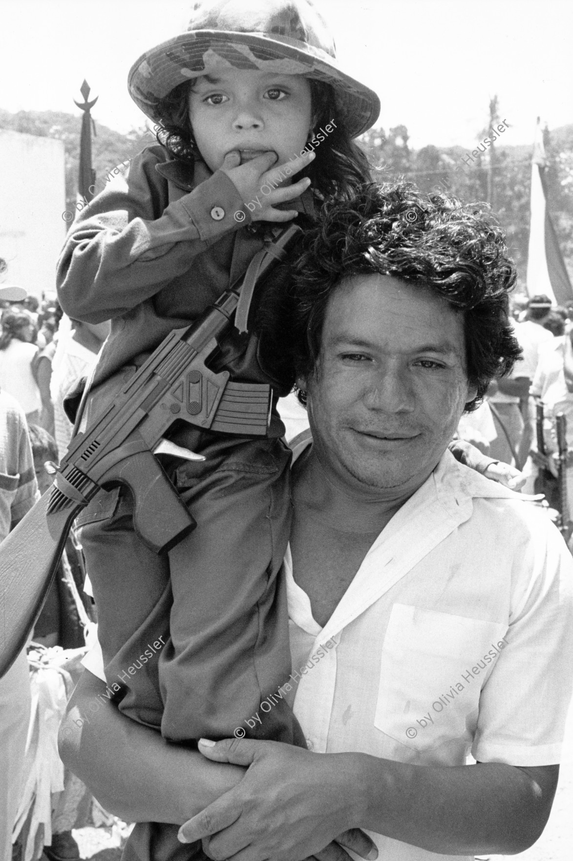 Image of sheet 19840290 photo 11: Geburtstag Sandinos. Vater trägt sein Mädchen in einer Uniform und Spielzeug Kalaschnikow AK-47 waffe aus plastic auf dem Arm. Des Generals des freien Menschen, wird gefeiert. «Sandino vive». Mascottchen kleines Kind in Uniform. Sandino  Nicaragua 1984 mascota child baby military sandinist Central america FSLN uniform girl rifle machine gun war arms father hut cachorro √