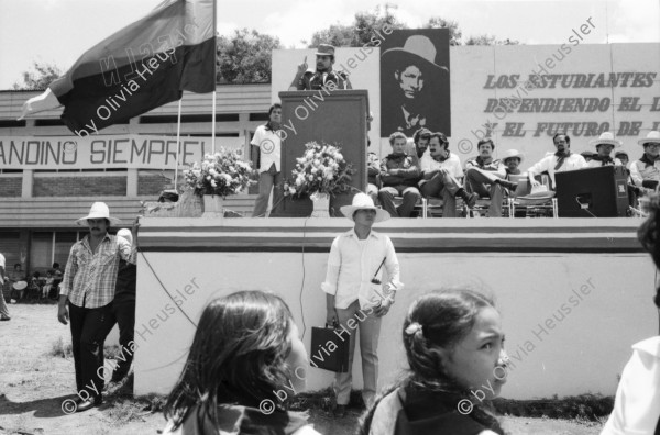 Image of sheet 19840290 photo 21: Geburtstag Sandinos. Des Generals des freien Menschen, wird gefeiert. «Sandino vive» steht an den Mauern. Kinder in Reihe. Gruppenbild. Bühne. FSLN Fahne. Mann Soldat mit Maske. Kommandanten mit Strohhüten. Fahnen. Mascottchen kleines Kind in Uniform. Marimbas werden an der Demo für Sandino mitgetragen. Masken werden getragen. Mural Wandmalerei Ein junger Soldat der den Helikopter abgeschossen hat mit den Honduranern drin. Nicaragua 1984