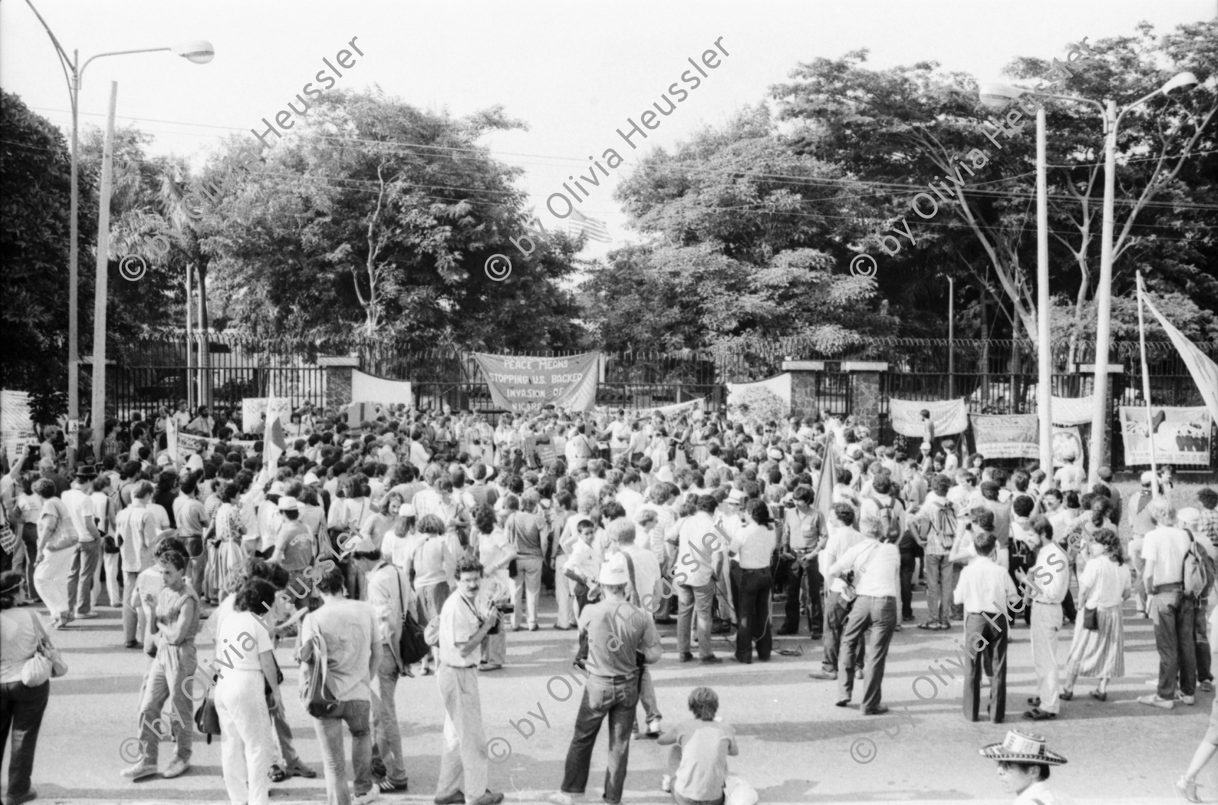 Image of sheet 19840410 photo 21: Viele Nordamerikaner Innen demonstrieren vor ihrer Botschaft. Protest US Amerikaner innen. U.S.A. Portrai Frau mit hut. Die Fahne weht auch stolz über ihr. Peace means stoping US backed Invasion of Nicaragua. Marsch am 19.Juli auf der Carretera Norte, vorbei an den Texnicsa. Wandbild mit Sandino.