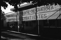 Image of sheet 19840420 photo 0: Todo para los frente de Guerra. Todo para los Combatientes. Am 5. Jahrestag der Revolution festen auch die Fabriken an der Carretera Norte. Fabrikarbeiter. Sicht durch die Ruinen aufs Gebäude der Banco de America. Im Vordergrund Demonstration. Die Fahnen wehen für die Revolution auf dem Platz. Das Nic. Rote Kreuz verteilt den erhitzten Köpfen Trinkwasser. Erdbebenruinen. Managua 1984