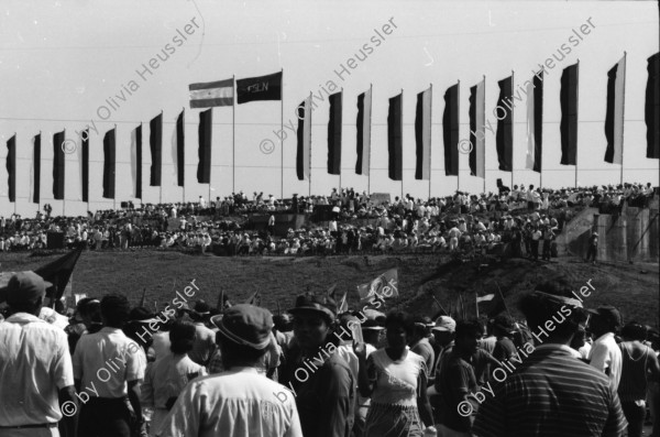 Image of sheet 19840420 photo 12: Todo para los frente de Guerra. Todo para los Combatientes. Am 5. Jahrestag der Revolution festen auch die Fabriken an der Carretera Norte. Fabrikarbeiter. Sicht durch die Ruinen aufs Gebäude der Banco de America. Im Vordergrund Demonstration. Die Fahnen wehen für die Revolution auf dem Platz. Das Nic. Rote Kreuz verteilt den erhitzten Köpfen Trinkwasser. Erdbebenruinen. Managua 1984