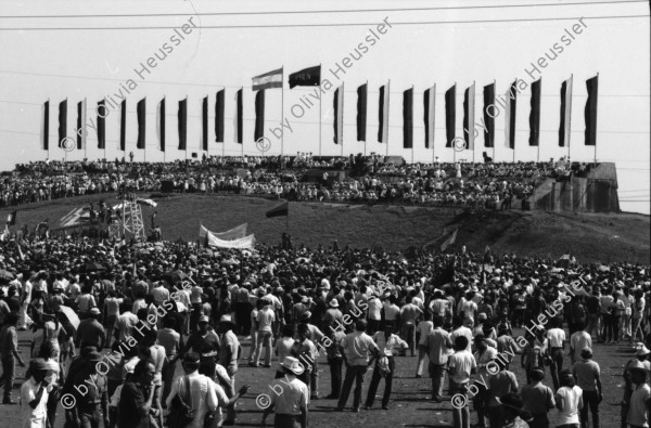 Image of sheet 19840420 photo 14: Todo para los frente de Guerra. Todo para los Combatientes. Am 5. Jahrestag der Revolution festen auch die Fabriken an der Carretera Norte. Fabrikarbeiter. Sicht durch die Ruinen aufs Gebäude der Banco de America. Im Vordergrund Demonstration. Die Fahnen wehen für die Revolution auf dem Platz. Das Nic. Rote Kreuz verteilt den erhitzten Köpfen Trinkwasser. Erdbebenruinen. Managua 1984