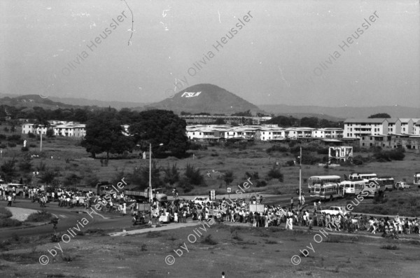 Image of sheet 19840420 photo 6: Todo para los frente de Guerra. Todo para los Combatientes. Am 5. Jahrestag der Revolution festen auch die Fabriken an der Carretera Norte. Fabrikarbeiter. Sicht durch die Ruinen aufs Gebäude der Banco de America. Im Vordergrund Demonstration. Die Fahnen wehen für die Revolution auf dem Platz. Das Nic. Rote Kreuz verteilt den erhitzten Köpfen Trinkwasser. Erdbebenruinen. Managua 1984