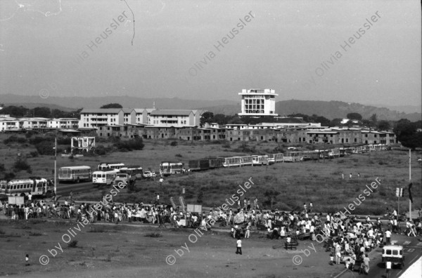 Image of sheet 19840420 photo 7: Todo para los frente de Guerra. Todo para los Combatientes. Am 5. Jahrestag der Revolution festen auch die Fabriken an der Carretera Norte. Fabrikarbeiter. Sicht durch die Ruinen aufs Gebäude der Banco de America. Im Vordergrund Demonstration. Die Fahnen wehen für die Revolution auf dem Platz. Das Nic. Rote Kreuz verteilt den erhitzten Köpfen Trinkwasser. Erdbebenruinen. Managua 1984