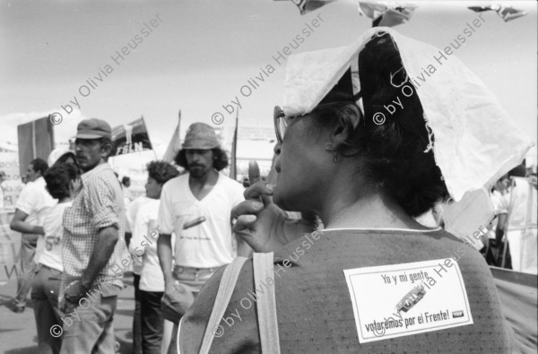 Image of sheet 19840610 photo 15: Nordamerikaner Innen unterstützen sandinistische Bauern der FSLN und der U.N.A.G. US-Amerikanische und Sandinistische Fahnen Flaggen nebeneinander. Frau in Kutsche grüsst die Kinder auf der Strasse. Mann auf Pferd. Mann als Frau verkleidet. Kinder verkleidet als enano auf der Plaza de Gobierno. Banco de America und im vordergrund Demonstration. Bauern mit Masken und Pauke. Managua Nicaragua 1984
 el enano cabezón .