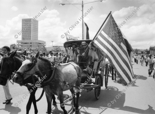 Image of sheet 19840610 photo 44: Nordamerikaner Innen unterstützen sandinistische Bauern der FSLN und der U.N.A.G. US-Amerikanische und Sandinistische Fahnen Flaggen nebeneinander. Banco de America und im vordergrund Demonstration. Bauern mit Masken und Pauke. Managua Nicaragua 1984