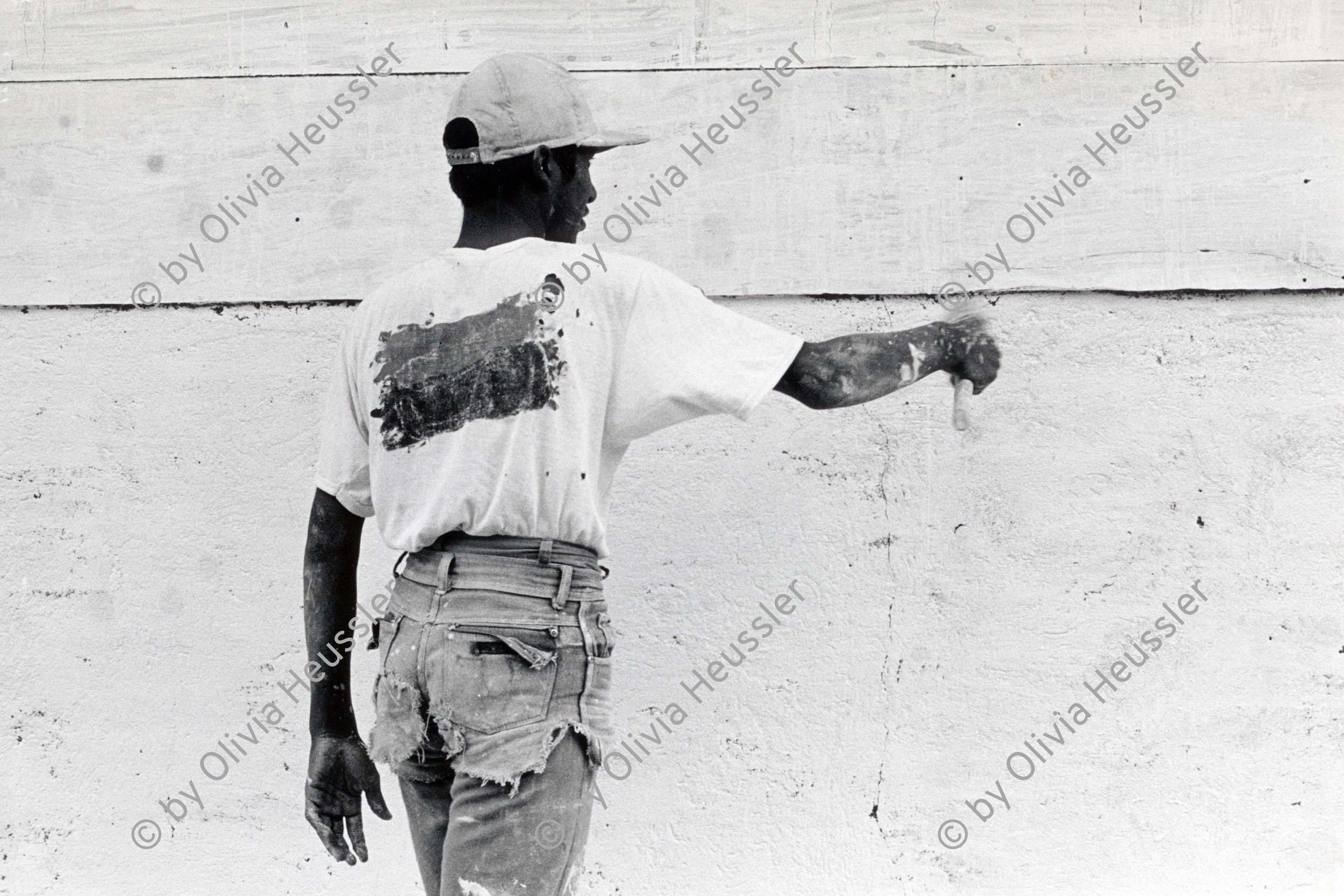 Image of sheet 19850370 photo 17: Ein junger Mann in abgeschnittenen Jeans bepinselt bemalt sein Haus mit weisser Farbe. Auf seinem Rücken trägt er die Farben Rot und Schwarz. Sie sind die Propagandafarben für die Sandinisten FSLN zu den Wahlen. San Juan del Sur Nicaragua 1985 √