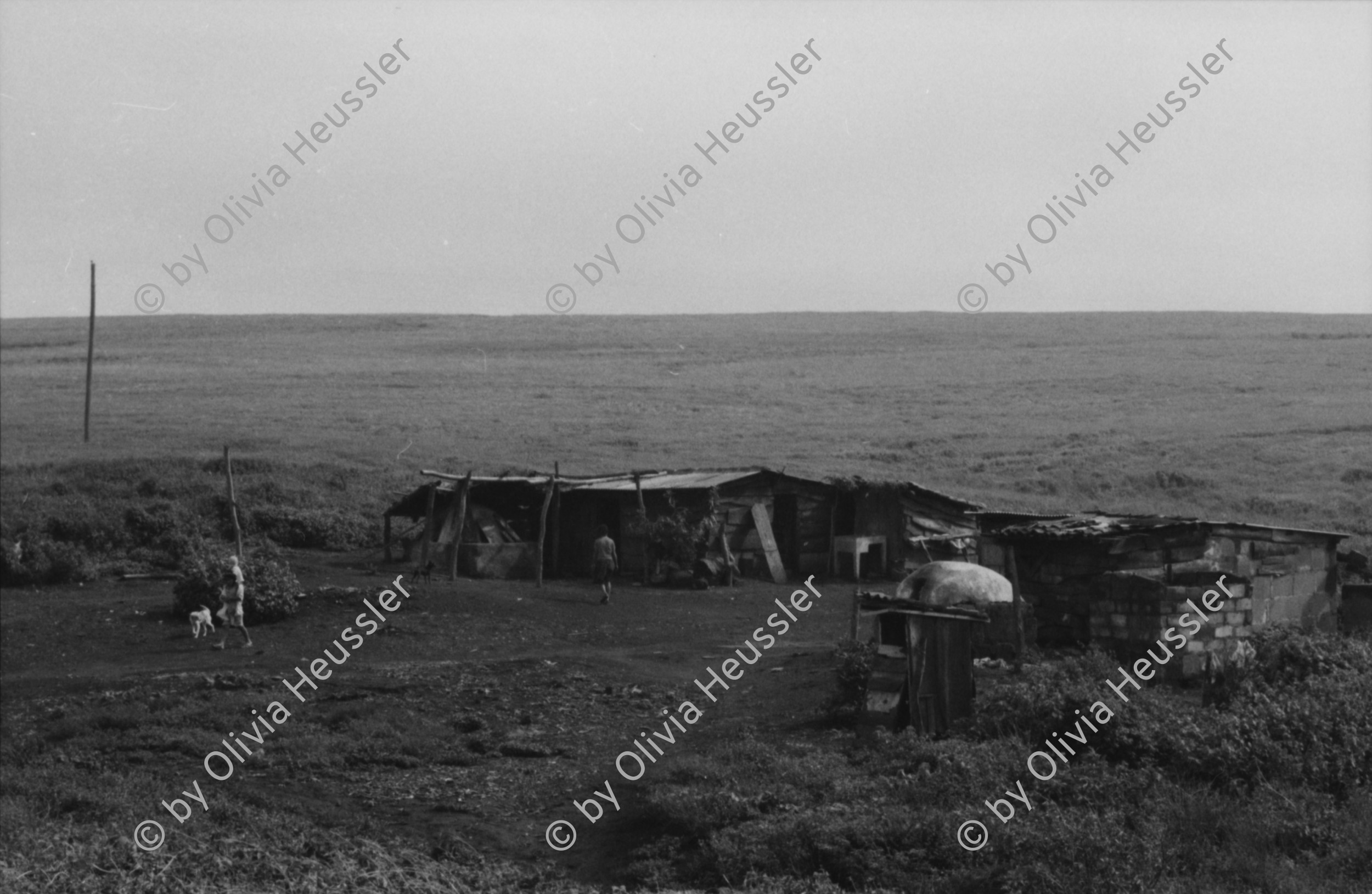 Image of sheet 19850440 photo 20: Am frühen Morgen fahre ich nach El Crucero . Es liegt hoch über Managua und es liegt immer im Nebel des Schwefeldampfes vom Vulkan Masaya. es ist ein paar Grade kühler dort. Der Schwefeldampf vom Vulkan in  Masaya, dem Santiago macht es der Natur unmöglich zu gedeihen. Bäume und andere Pflanzen ertragen
diese Luft nicht. Trotzdem stehen da einige Häuser und vereinzelt sehe ich Menschen. Frau mit Rind Kinder stehen da. Ein Mädchen vor einer Bretterwand Haus. Wandmalereien. COSEP Leute an PK.
José Chepe auf Motorrad Trautes. Gruppenbild Carretera sur. Managua 1985