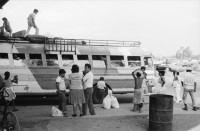 Image of sheet 19870061 photo 1: Bushaltestelle am Mercado R. Huembes. Menschen schleppen und bepacken die Busse. Der Buss fährt nach Masaya. Leute warten auf Buss, ich Olivia heussler spiegle mich im Spiegel des Wartenden. Menschenschlange wartet für den Buss. Managua Nicaragua 1987
