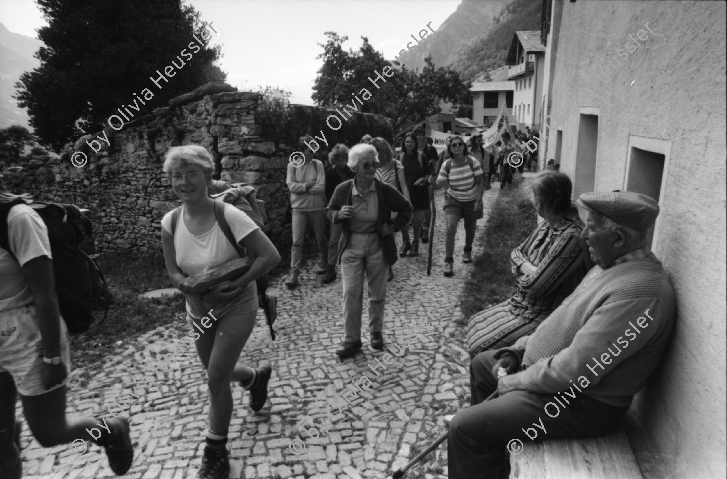 Image of sheet 19870251 photo 33: Frauen Protest Wanderung gegen den Plan eines Pumpspeicherkraftwerks im Val Madris Passüberquerung auf alter Rinder Treppe Kuhtreppe für Kühe ins Bergell. Graubünden Einmarsch ins Dorf Soglio.  Rentner Zuschauer sitzen im Dorf. Transparent «Wir Frauen haben genug Energie»
Bergell Soglio Graubünden Schweiz 1987
