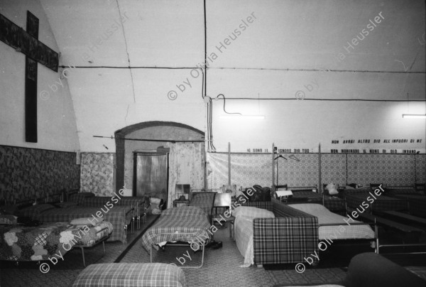 Image of sheet 19870390 photo 32: Im Auffanglager für die von der Schweiz abgewiesenen Flüchtlinge und Obdachlose unter den Geleisen des Zentral Bahnhofs, Mailand 1987. Bei Fratello Ettore der Raum dient auch als Kirche, Schlaf- und Essraum. Bevor die vorwiegend aus Nordafrika kommenden islamischen Männer ihre Suppe kriegen, müssen sie sich die christliche Messe anhören. Viele verdecken ihre Gesichter vor der Camera und mir. Betten stehen da vor einem Bild eines Heiligen der auch liegt, nur nicht so bequem. Mann bittet vor Einlass in die Katakomben. Milano Italien Italy Mailand 1987