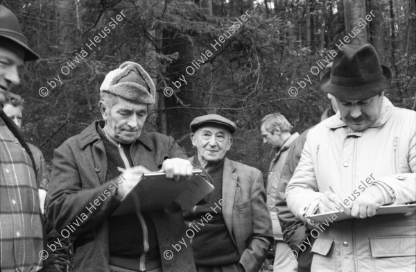 Image of sheet 19880040 photo 1: alter Männer stehen zusammen an Holzgant.Sie schreiben sich die Preise auf. Percy Heussler ist auch dabei. Nr. 12 und 13. Die langen Holz Stiehle zum Cervelats Braten liegen bereit. Die Männer braten Cervelats über dem Feuer. Ein alter Mann zeigt in die Luft. Die Cervelats liegen zum Braten bereit. Die Männer geniessen etwas warmes in der winterlichen Kälte. (!) Holzgant Holzverarbeitung Fällanden Kanton Zürich Schweiz wood forest market man men meat sausage