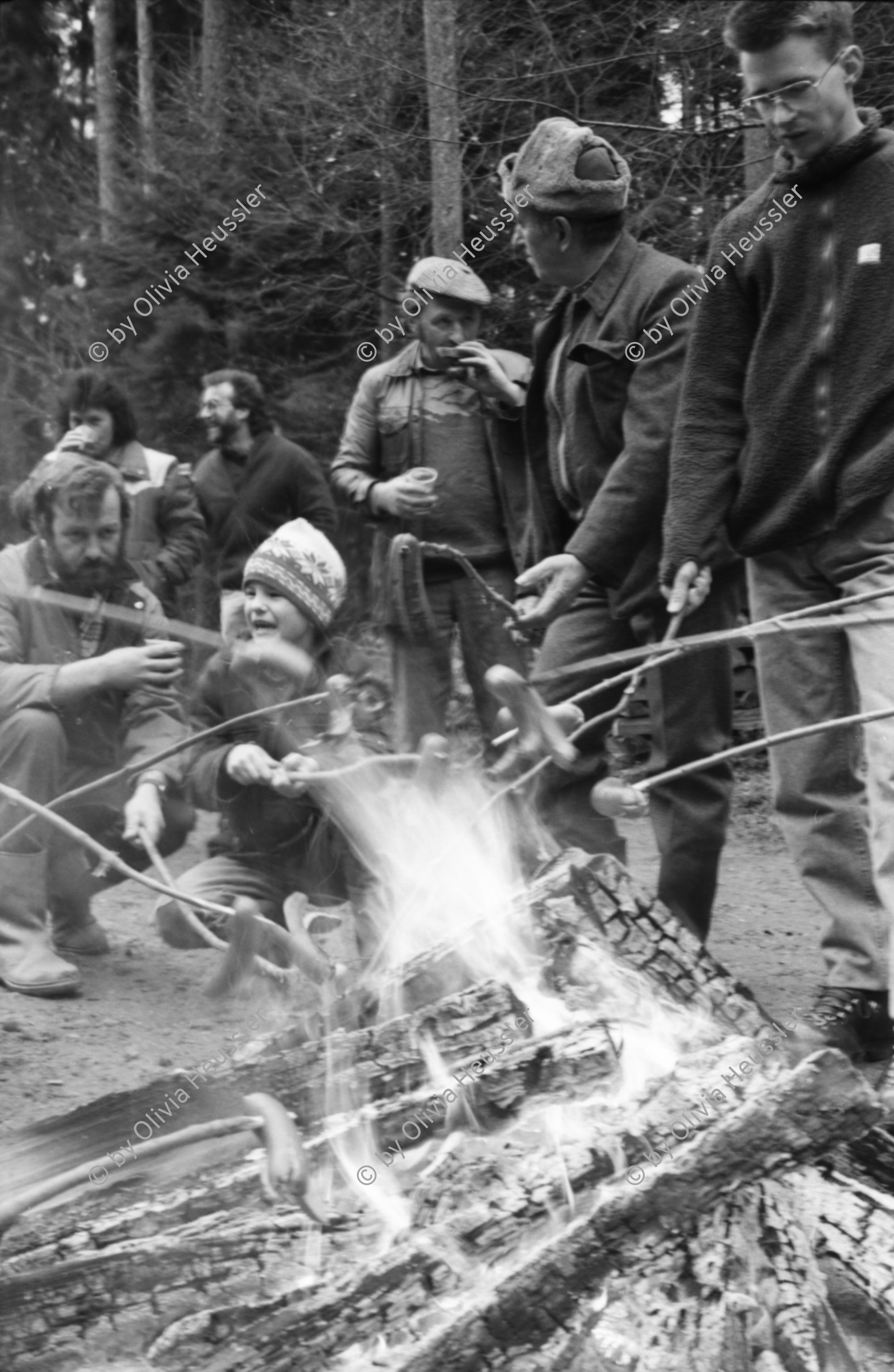 Image of sheet 19880040 photo 21: alter Männer stehen zusammen an Holzgant.Sie schreiben sich die Preise auf. Percy Heussler ist auch dabei. Nr. 12 und 13. Die langen Holz Stiehle zum Cervelats Braten liegen bereit. Die Männer braten Cervelats über dem Feuer. Ein alter Mann zeigt in die Luft. Die Cervelats liegen zum Braten bereit. Die Männer geniessen etwas warmes in der winterlichen Kälte. (!) Holzgant Holzverarbeitung Fällanden Kanton Zürich Schweiz wood forest market man men meat sausage
