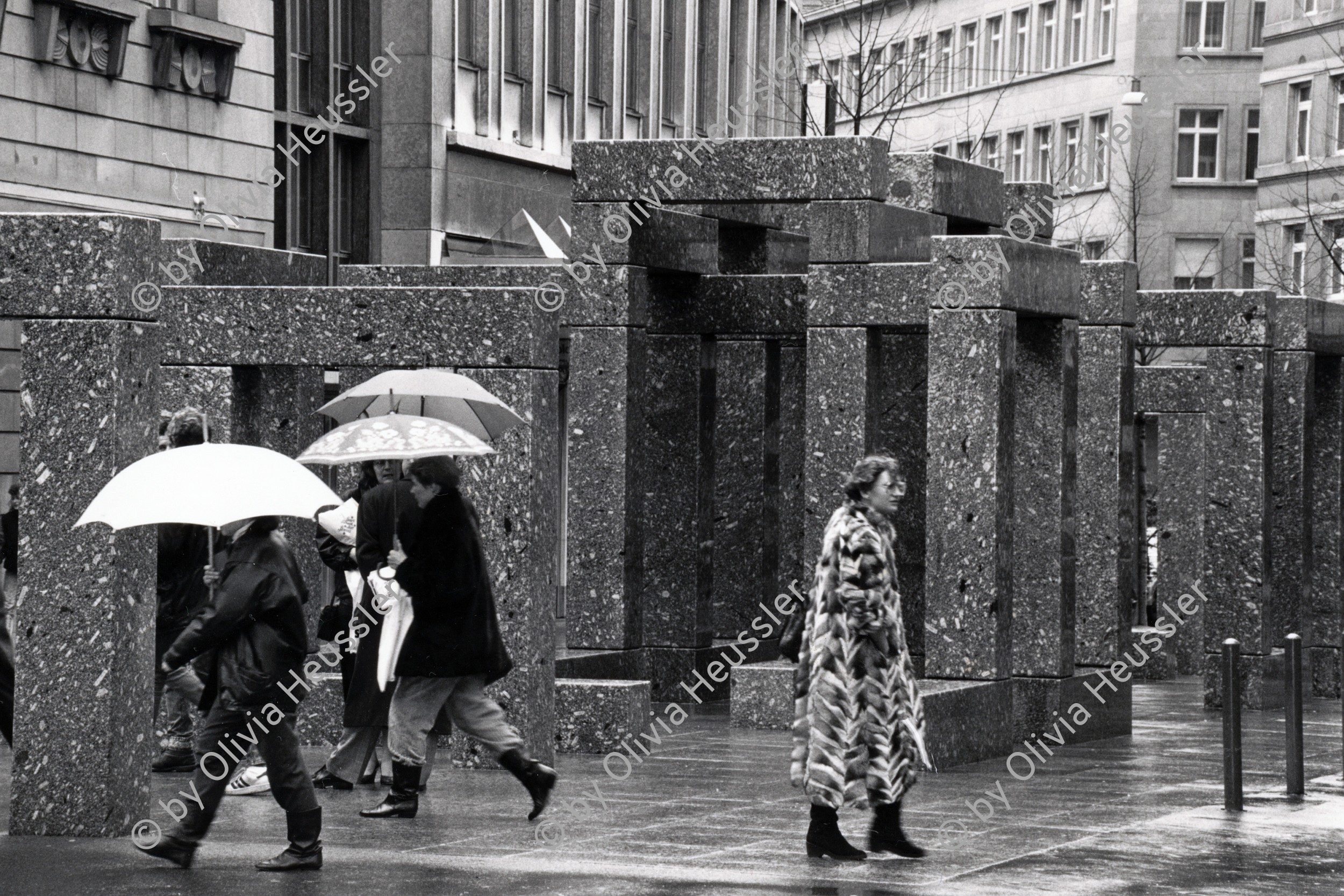 Image of sheet 19880250 photo 35: Szene an der Bahnhofstrasse. Frauen in Pelzmäntel unter Regenschirmen mit Hunden 
Granitskulptur (1983) von Max Bill
Der Schweizer Künstler Max Bill schuf 1983 die begehbare «Pavillon-Skulptur» – ein Beispiel für klare Linien –, die heute an erstklassiger Lage vor dem Zürcher Hauptsitz der Grossbank UBS an der Bahnhofstrasse steht. Die streng konzipierte, von Logik und Klarheit geprägte Konstruktion aus hochpolierten, grauen Granitelementen war anfänglich äusserst umstritten.
Bahnhofstrasse 
Zürich 1988 √