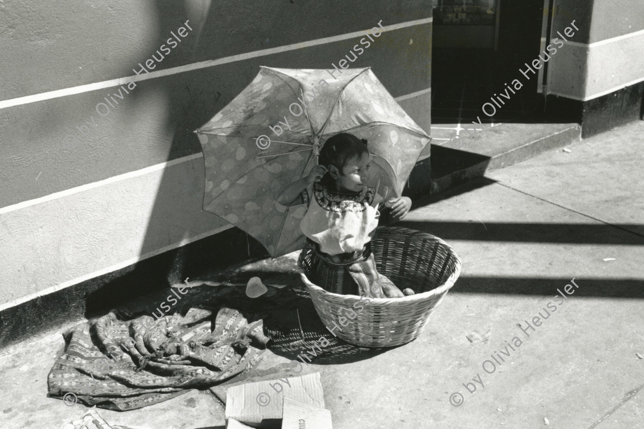 Image of sheet 19880990 photo 21: © 1988, Olivia Heussler/www.clic.li
Kinderarbeit 
Guatemala City Stadt kleines Mädchen sitzt  am Markt in einem Korb unter einem Regenschirm Sonnenschirm 1988 √
girl work street market