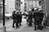 Image of sheet 19890330 photo 5: frtsg. 890320        Ein Kommandant der Zürcher Stadtpolizei entfernt an der Hausfassade der besetzten Köchlistrasse 22 (18.3.-6.4.89) ein Transparent. Grenadiere und Spitzel ! räumen das Haus. Ein Grendadier bestückt mit einem Korbschild sitzt auf