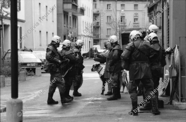 Image of sheet 19890330 photo 5: frtsg. 890320        Ein Kommandant der Zürcher Stadtpolizei entfernt an der Hausfassade der besetzten Köchlistrasse 22 (18.3.-6.4.89) ein Transparent. Grenadiere und Spitzel ! räumen das Haus. Ein Grendadier bestückt mit einem Korbschild sitzt auf