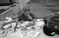 Image of sheet 19900730 photo 13: Am Checkpoint Charlie in Berlin verkaufen Polen alte DDR Uniformen und Flaggen etc. Im Hintergrund steht 'Neue Zeit'. Verkäufer ist uniformiert. Alter Mann hinter den Mützen. 
Former Checkpoint Wallborder to East-Germany GDR. An old Polish Citizen is sitting near old military uniforms from various Eastern Countries. The Pole is selling these uniforms to travellers who visit whatever is left over from the 'Wall'.

Potsdamer-Platz ist besetzt von Obdachlosen in Zelten. Refugees from the Eastern block are sitting in front of their tents on the Potsdamerplatz near the former wall that divided East- and Westgermany. Trabant fährt auf der Ebertstrasse auf ehemaligem Mauerstreifen. Brandenburger Tor Brandenburg Gate ohne Skulptur Quadriga. Frau fotografiert. Schwarzmarkt. Polenmarkt. Volks- Polizei bricht illegale Verkaufsstände ab. Alte kontrolliert ihre Kasse Portemonnaie in Pankov. Deutschland 1990

Checkpoint Charlie (or Checkpoint C) was the name given by the Western Allies to the best-known Berlin Wall crossing point between East Berlin and West Berlin during the Cold War.

The Soviet Union prompted the erection of the Berlin Wall in 1961 to stem the flow of Eastern Bloc emigration westward through what had become a loophole in the Soviet border system, preventing escape over the city sector border from East Berlin to West Berlin. Checkpoint Charlie became a symbol of the Cold War, representing the separation of east and west, and—for some East Germans—a gateway to freedom. Soviet and American tanks briefly faced off at the location during the Berlin Crisis of 1961.