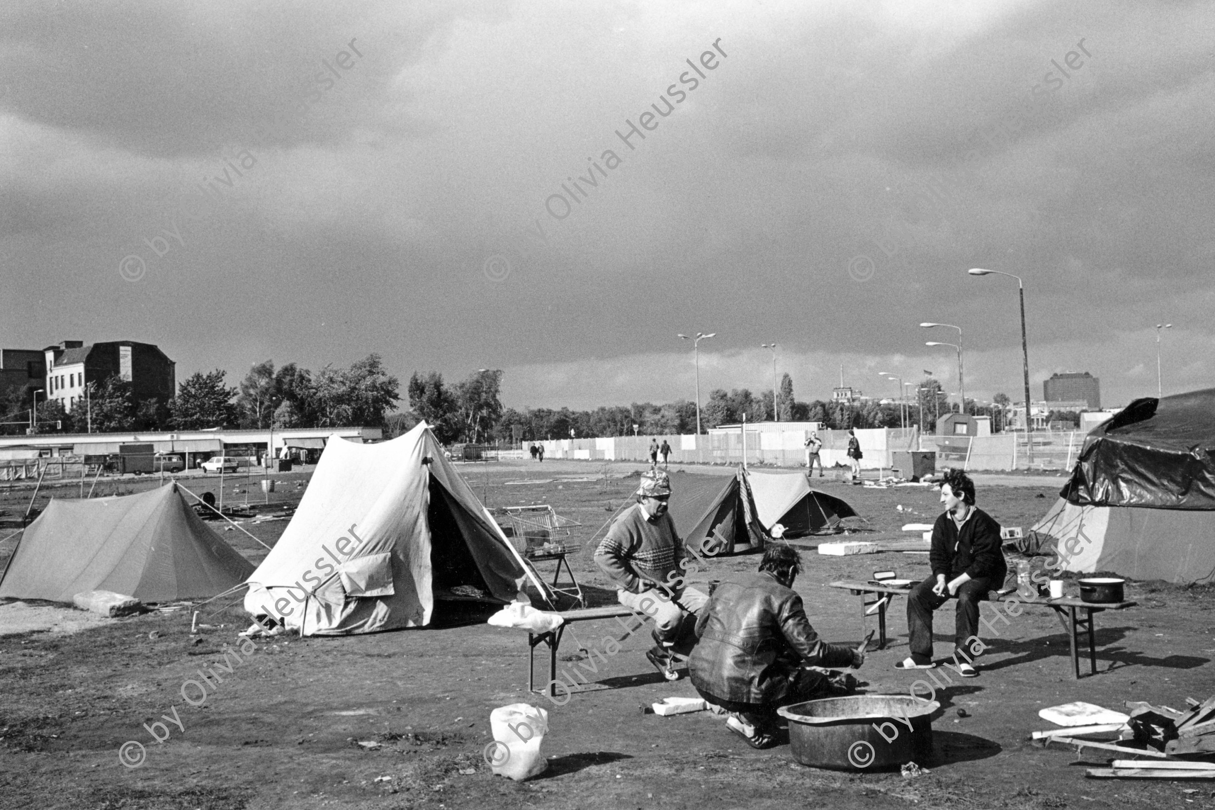 Image of sheet 19900730 photo 19: © 1990, Olivia Heussler / www.clic.li
Potsdamer-Platz ist besetzt von Obdachlosen in Zelten. Refugees from the Eastern block are sitting in front of their tents on the Potsdamerplatz near the former wall that divided East- and Westgermany. Ebertstrasse auf ehemaligem Mauerstreifen. Deutschland 1990 √ Berlin

Potsdamer Platz (German: [potsdamaː plats] ( listen), literally Potsdam Square) is an important public square and traffic intersection in the centre of Berlin, Germany, lying about one kilometre south of the Brandenburg Gate and the Reichstag (German Parliament Building), and close to the southeast corner of the Tiergarten park. It is named after the city of Potsdam, some 25 km to the south west, and marks the point where the old road from Potsdam passed through the city wall of Berlin at the Potsdam Gate. After developing within the space of little over a century from an intersection of rural thoroughfares into the most bustling traffic intersection in Europe it was totally laid waste during World War II and then left desolate during the Cold War era when the Berlin Wall bisected its former location. Since German reunification, Potsdamer Platz has been the site of major redevelopment projects.