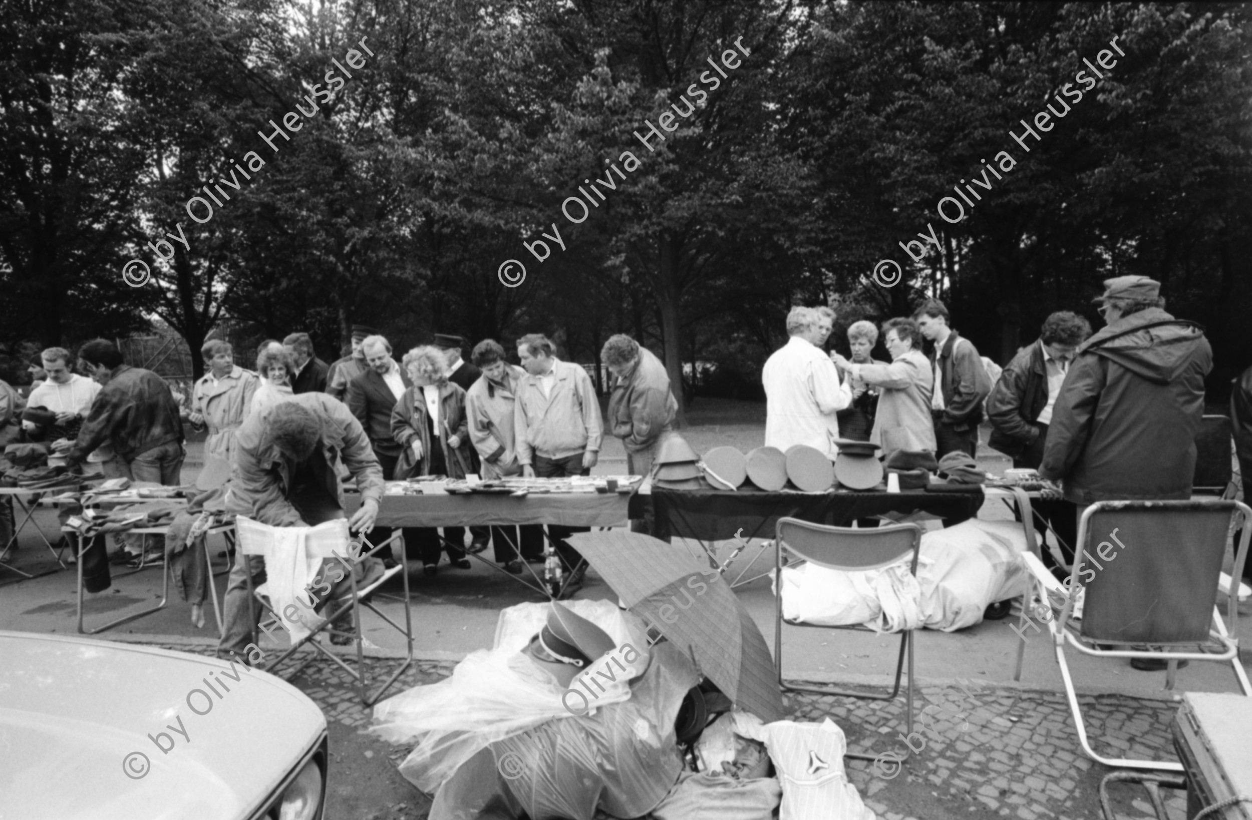 Image of sheet 19900730 photo 26: Am Checkpoint Charlie in Berlin verkaufen Polen alte DDR Uniformen und Flaggen etc. Im Hintergrund steht 'Neue Zeit'. Verkäufer ist uniformiert. Alter Mann hinter den Mützen. 
Former Checkpoint Wallborder to East-Germany GDR. An old Polish Citizen is sitting near old military uniforms from various Eastern Countries. The Pole is selling these uniforms to travellers who visit whatever is left over from the 'Wall'.

Potsdamer-Platz ist besetzt von Obdachlosen in Zelten. Refugees from the Eastern block are sitting in front of their tents on the Potsdamerplatz near the former wall that divided East- and Westgermany. Trabant fährt auf der Ebertstrasse auf ehemaligem Mauerstreifen. Brandenburger Tor Brandenburg Gate ohne Skulptur Quadriga. Frau fotografiert. Schwarzmarkt. Polenmarkt. Volks- Polizei bricht illegale Verkaufsstände ab. Alte kontrolliert ihre Kasse Portemonnaie in Pankov. Deutschland 1990

Checkpoint Charlie (or Checkpoint C) was the name given by the Western Allies to the best-known Berlin Wall crossing point between East Berlin and West Berlin during the Cold War.

The Soviet Union prompted the erection of the Berlin Wall in 1961 to stem the flow of Eastern Bloc emigration westward through what had become a loophole in the Soviet border system, preventing escape over the city sector border from East Berlin to West Berlin. Checkpoint Charlie became a symbol of the Cold War, representing the separation of east and west, and—for some East Germans—a gateway to freedom. Soviet and American tanks briefly faced off at the location during the Berlin Crisis of 1961.