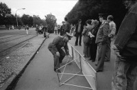 Image of sheet 19900730 photo 28: Am Checkpoint Charlie in Berlin verkaufen Polen alte DDR Uniformen und Flaggen etc. Im Hintergrund steht 'Neue Zeit'. Verkäufer ist uniformiert. Alter Mann hinter den Mützen. 
Former Checkpoint Wallborder to East-Germany GDR. An old Polish Citizen is sitting near old military uniforms from various Eastern Countries. The Pole is selling these uniforms to travellers who visit whatever is left over from the 'Wall'.

Potsdamer-Platz ist besetzt von Obdachlosen in Zelten. Refugees from the Eastern block are sitting in front of their tents on the Potsdamerplatz near the former wall that divided East- and Westgermany. Trabant fährt auf der Ebertstrasse auf ehemaligem Mauerstreifen. Brandenburger Tor Brandenburg Gate ohne Skulptur Quadriga. Frau fotografiert. Schwarzmarkt. Polenmarkt. Volks- Polizei bricht illegale Verkaufsstände ab. Alte kontrolliert ihre Kasse Portemonnaie in Pankov. Deutschland 1990

Checkpoint Charlie (or Checkpoint C) was the name given by the Western Allies to the best-known Berlin Wall crossing point between East Berlin and West Berlin during the Cold War.

The Soviet Union prompted the erection of the Berlin Wall in 1961 to stem the flow of Eastern Bloc emigration westward through what had become a loophole in the Soviet border system, preventing escape over the city sector border from East Berlin to West Berlin. Checkpoint Charlie became a symbol of the Cold War, representing the separation of east and west, and—for some East Germans—a gateway to freedom. Soviet and American tanks briefly faced off at the location during the Berlin Crisis of 1961.