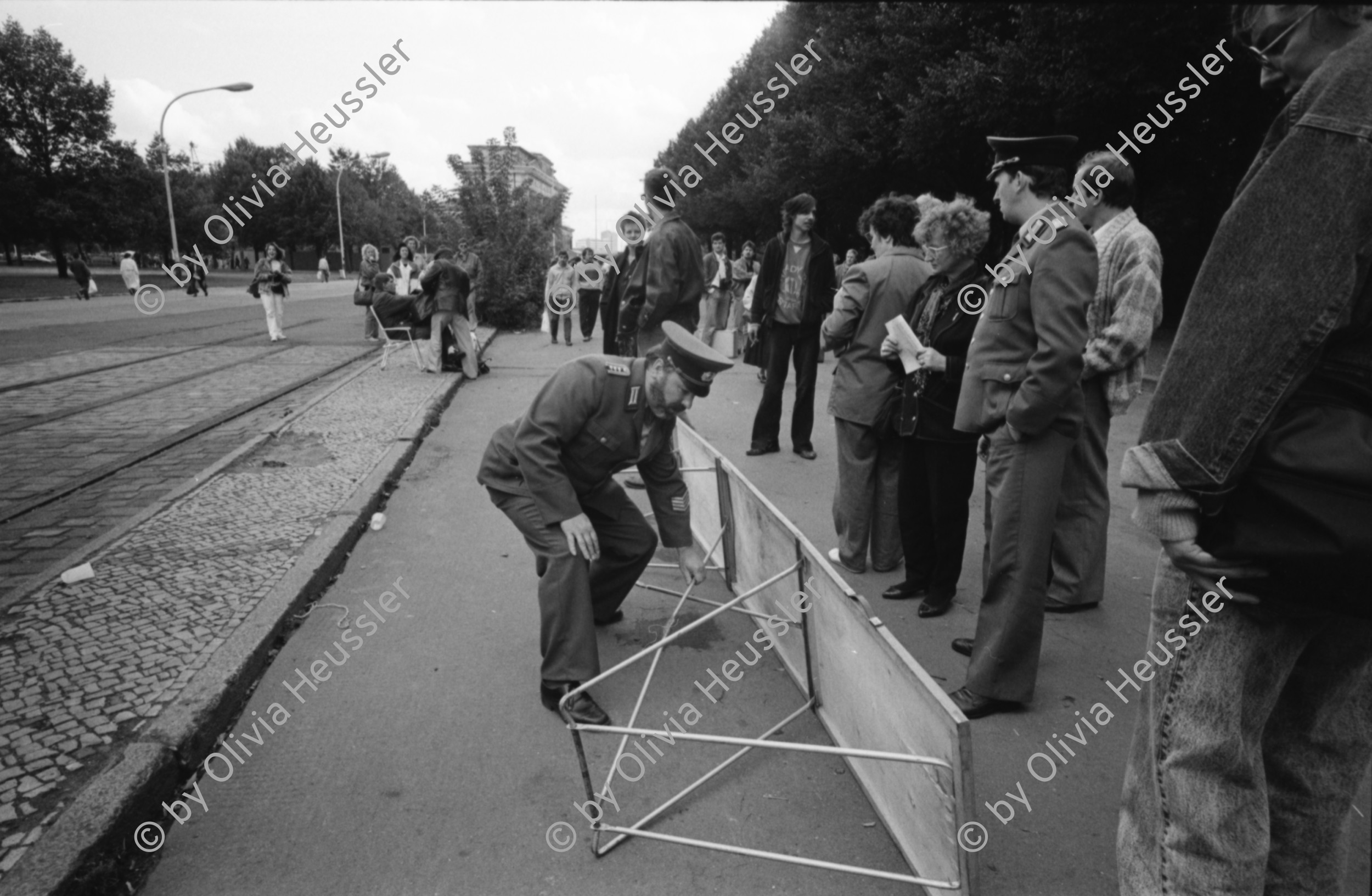 Image of sheet 19900730 photo 28: Am Checkpoint Charlie in Berlin verkaufen Polen alte DDR Uniformen und Flaggen etc. Im Hintergrund steht 'Neue Zeit'. Verkäufer ist uniformiert. Alter Mann hinter den Mützen. 
Former Checkpoint Wallborder to East-Germany GDR. An old Polish Citizen is sitting near old military uniforms from various Eastern Countries. The Pole is selling these uniforms to travellers who visit whatever is left over from the 'Wall'.

Potsdamer-Platz ist besetzt von Obdachlosen in Zelten. Refugees from the Eastern block are sitting in front of their tents on the Potsdamerplatz near the former wall that divided East- and Westgermany. Trabant fährt auf der Ebertstrasse auf ehemaligem Mauerstreifen. Brandenburger Tor Brandenburg Gate ohne Skulptur Quadriga. Frau fotografiert. Schwarzmarkt. Polenmarkt. Volks- Polizei bricht illegale Verkaufsstände ab. Alte kontrolliert ihre Kasse Portemonnaie in Pankov. Deutschland 1990

Checkpoint Charlie (or Checkpoint C) was the name given by the Western Allies to the best-known Berlin Wall crossing point between East Berlin and West Berlin during the Cold War.

The Soviet Union prompted the erection of the Berlin Wall in 1961 to stem the flow of Eastern Bloc emigration westward through what had become a loophole in the Soviet border system, preventing escape over the city sector border from East Berlin to West Berlin. Checkpoint Charlie became a symbol of the Cold War, representing the separation of east and west, and—for some East Germans—a gateway to freedom. Soviet and American tanks briefly faced off at the location during the Berlin Crisis of 1961.
