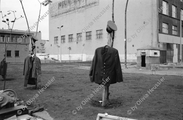 Image of sheet 19900730 photo 4: Am Checkpoint Charlie in Berlin verkaufen Polen alte DDR Uniformen und Flaggen etc. Im Hintergrund steht 'Neue Zeit'. Verkäufer ist uniformiert. Alter Mann hinter den Mützen. 
Former Checkpoint Wallborder to East-Germany GDR. An old Polish Citizen is sitting near old military uniforms from various Eastern Countries. The Pole is selling these uniforms to travellers who visit whatever is left over from the 'Wall'.

Potsdamer-Platz ist besetzt von Obdachlosen in Zelten. Refugees from the Eastern block are sitting in front of their tents on the Potsdamerplatz near the former wall that divided East- and Westgermany. Trabant fährt auf der Ebertstrasse auf ehemaligem Mauerstreifen. Brandenburger Tor Brandenburg Gate ohne Skulptur Quadriga. Frau fotografiert. Schwarzmarkt. Polenmarkt. Volks- Polizei bricht illegale Verkaufsstände ab. Alte kontrolliert ihre Kasse Portemonnaie in Pankov. Deutschland 1990

Checkpoint Charlie (or Checkpoint C) was the name given by the Western Allies to the best-known Berlin Wall crossing point between East Berlin and West Berlin during the Cold War.

The Soviet Union prompted the erection of the Berlin Wall in 1961 to stem the flow of Eastern Bloc emigration westward through what had become a loophole in the Soviet border system, preventing escape over the city sector border from East Berlin to West Berlin. Checkpoint Charlie became a symbol of the Cold War, representing the separation of east and west, and—for some East Germans—a gateway to freedom. Soviet and American tanks briefly faced off at the location during the Berlin Crisis of 1961.