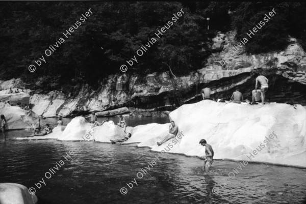 Image of sheet 19910570 photo 11: Flussbaden in der Melezza im Tessiner Centovalli ist immer mehr beliebt. Das von der Sonne gewärmte Felsgestein ist nach dem kalten Wasser eine Heizung. Die Frauen liegen bequem in den Nischen. Tegna the Best. Auf einem Frottier tuch ist eine  Frau abgebildet. Gummiboot und Männerschulter. Sandstrand  Männer sitzen in Badehosen auf den hohen Felsen. Ein ausgestopftes Krokodil (?) liegt hinter dem Heckfenster im Auto Fahrzeug. Zeitung lesend in der Hängematte. Tessin Ticino Pozzo 1991 Tourismus Baden Sonnen Bikini