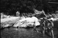 Image of sheet 19910570 photo 18: Flussbaden in der Melezza im Tessiner Centovalli ist immer mehr beliebt. Das von der Sonne gewärmte Felsgestein ist nach dem kalten Wasser eine Heizung. Die Frauen liegen bequem in den Nischen. Tegna the Best. Auf einem Frottier tuch ist eine  Frau abgebildet. Gummiboot und Männerschulter. Sandstrand  Männer sitzen in Badehosen auf den hohen Felsen. Ein ausgestopftes Krokodil (?) liegt hinter dem Heckfenster im Auto Fahrzeug. Zeitung lesend in der Hängematte. Tessin Ticino Pozzo 1991 Tourismus Baden Sonnen Bikini