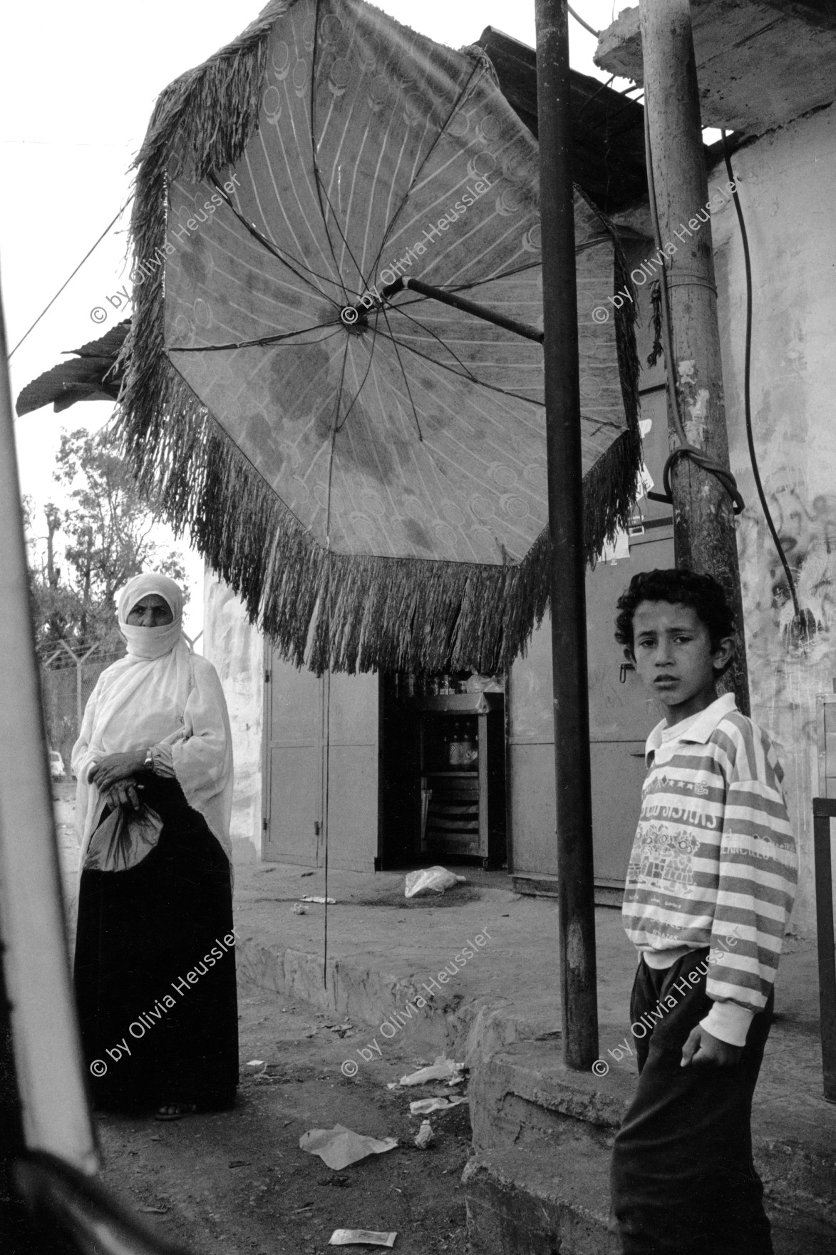 Image of sheet 19920300 photo 35: Auf dem Weg ins Jabalia Camp, Gaza 04.92 Palästina
On the way to Jabalia Camp, 1992  √ Palestine Gaza strip israeli occupied territory

© 1992, OLIVIA HEUSSLER / www.clic.li

Aus: Out of Jerusalem / Jenseits von Jerusalem page 93
A palestinian family in Jabalia refugee camp refugees boy child mother headscarf woman umbrella sun street 
Jabalia Camp (Arabic: مخيّم جباليا‎) is a Palestinian refugee camp located 3 kilometers (1.9 mi) north of Jabalia. The refugee camp is in the North Gaza Governorate, Gaza Strip. According to the Palestinian Central Bureau of Statistics, the camp had a population of 93,455 in mid-year 2006