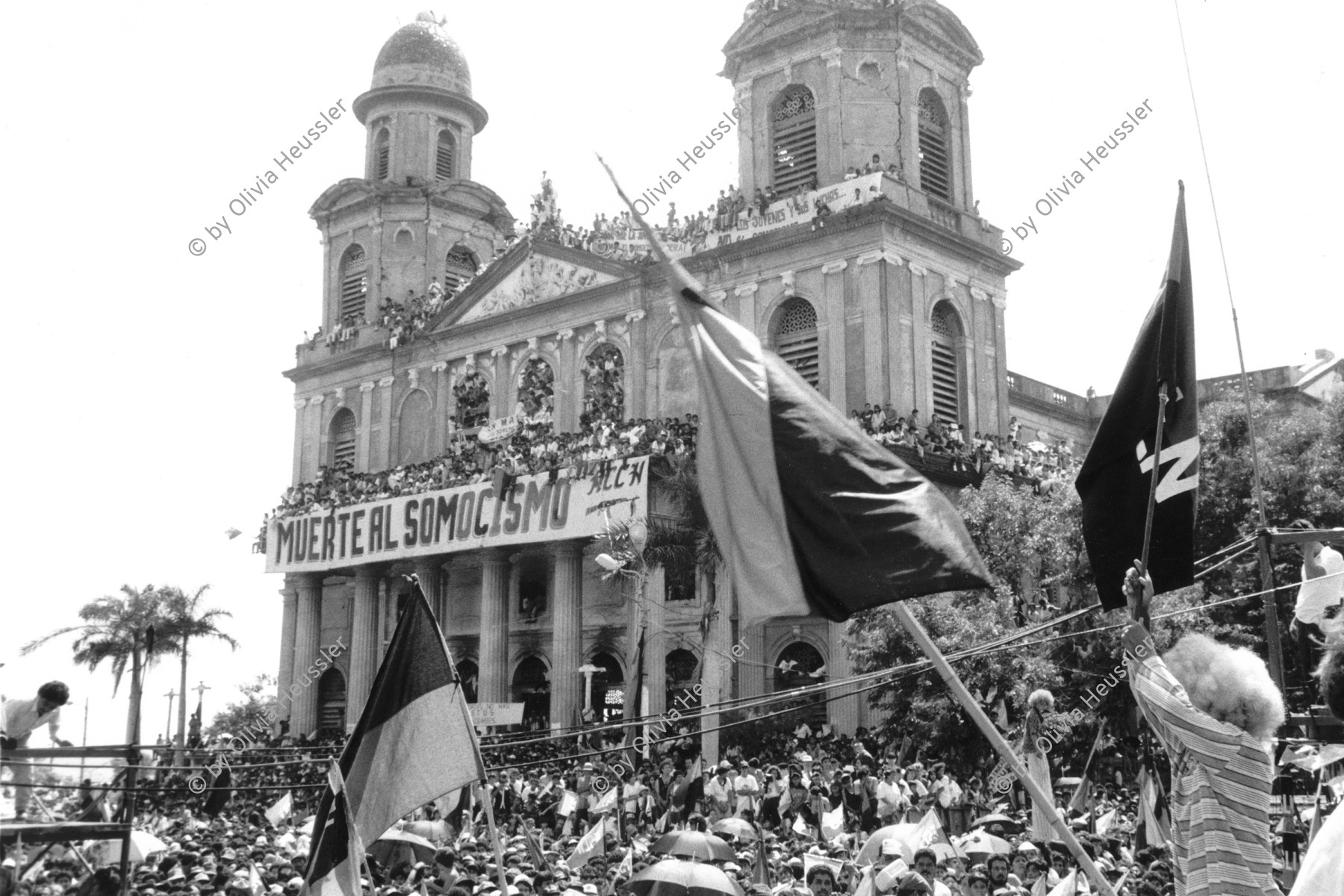 Image of sheet 19930380 photo 36: Menschenmassen Protest Demonstration Jubeln Feier am 19.7.93 auf der Plaza de la Revolucion, vor der Cathedrale Kathedrale: Managua ( 14. Jahrestag der sandinistischen Revolution ) FSLN Nicaragua 1993 √ 19. Juli 1993
'Muerte al Somocismo' Steht auf dem Banner. Sandinistische flaggen und Fahnen. Clown mit Perrücke

Durante las celebraciones del 14° aniversario
de la Revolución sandinista. Plaza de la Revolución, Managua, 19 de julio de 1993
Celebrating the 14th anniversary
of the Sandinista revolution.
Plaza of the Revolution, Managua, 1993
Die Kathedrale Santiago de Managua ist die Ruine der alten Kathedrale von Managua. Sie befindet sich an der Plaza de la Revolution, vormals Plaza de la República. Sie wurde als kaschierter Stahlskelettbau im neoklassischen Stil errichtet. Die Fassadenaufteilung ist ähnlich der von St-Sulpice de Paris.