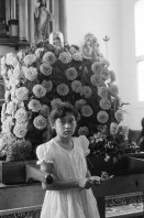 Image of sheet 19930520 photo 12: La vela de Santo Domingo, Iglesia de las Sierritas 9 de agosto 1993. Eine Schwester sitzt im Hintergrund.Sie überwacht aus Distanz die Feierlichkeiten die Tage später zu Toten führt , die in der Kirche ersticken. Der Dominguito ist ein kleiner Heiler und Heiliger unter einer Glashaube. Rausgeputzte Frau vor dem Blumenberg des Santos. Sie empfängt die Almosen und verteilt Blumen. Leute kommen auf den Knien zum Santos. Sie werden von einem mit Oel getränkten Mann geführt. Andenken und Glücksbringer werden verkauft. Managua Nicaragua 1993