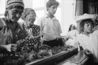 Image of sheet 19930520 photo 14: La vela de Santo Domingo, Iglesia de las Sierritas 9 de agosto 1993. Eine Schwester sitzt im Hintergrund.Sie überwacht aus Distanz die Feierlichkeiten die Tage später zu Toten führt , die in der Kirche ersticken. Der Dominguito ist ein kleiner Heiler und Heiliger unter einer Glashaube. Rausgeputzte Frau vor dem Blumenberg des Santos. Sie empfängt die Almosen und verteilt Blumen. Leute kommen auf den Knien zum Santos. Sie werden von einem mit Oel getränkten Mann geführt. Andenken und Glücksbringer werden verkauft. Managua Nicaragua 1993