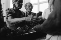 Image of sheet 19930520 photo 18: La vela de Santo Domingo, Iglesia de las Sierritas 9 de agosto 1993. Eine Schwester sitzt im Hintergrund.Sie überwacht aus Distanz die Feierlichkeiten die Tage später zu Toten führt , die in der Kirche ersticken. Der Dominguito ist ein kleiner Heiler und Heiliger unter einer Glashaube. Rausgeputzte Frau vor dem Blumenberg des Santos. Sie empfängt die Almosen und verteilt Blumen. Leute kommen auf den Knien zum Santos. Sie werden von einem mit Oel getränkten Mann geführt. Andenken und Glücksbringer werden verkauft. Managua Nicaragua 1993