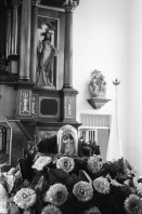 Image of sheet 19930520 photo 21: La vela de Santo Domingo, Iglesia de las Sierritas 9 de agosto 1993. Eine Schwester sitzt im Hintergrund.Sie überwacht aus Distanz die Feierlichkeiten die Tage später zu Toten führt , die in der Kirche ersticken. Der Dominguito ist ein kleiner Heiler und Heiliger unter einer Glashaube. Rausgeputzte Frau vor dem Blumenberg des Santos. Sie empfängt die Almosen und verteilt Blumen. Leute kommen auf den Knien zum Santos. Sie werden von einem mit Oel getränkten Mann geführt. Andenken und Glücksbringer werden verkauft. Managua Nicaragua 1993