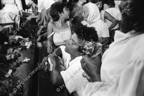 Image of sheet 19930520 photo 22: La vela de Santo Domingo, Iglesia de las Sierritas 9 de agosto 1993. Eine Schwester sitzt im Hintergrund.Sie überwacht aus Distanz die Feierlichkeiten die Tage später zu Toten führt , die in der Kirche ersticken. Der Dominguito ist ein kleiner Heiler und Heiliger unter einer Glashaube. Rausgeputzte Frau vor dem Blumenberg des Santos. Sie empfängt die Almosen und verteilt Blumen. Leute kommen auf den Knien zum Santos. Sie werden von einem mit Oel getränkten Mann geführt. Andenken und Glücksbringer werden verkauft. Managua Nicaragua 1993