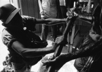 Image of sheet 19930520 photo 28: La vela de Santo Domingo, Iglesia de las Sierritas 9 de agosto 1993. Eine Schwester sitzt im Hintergrund.Sie überwacht aus Distanz die Feierlichkeiten die Tage später zu Toten führt , die in der Kirche ersticken. Der Dominguito ist ein kleiner Heiler und Heiliger unter einer Glashaube. Rausgeputzte Frau vor dem Blumenberg des Santos. Sie empfängt die Almosen und verteilt Blumen. Leute kommen auf den Knien zum Santos. Sie werden von einem mit Oel getränkten Mann geführt. Andenken und Glücksbringer werden verkauft. Managua Nicaragua 1993