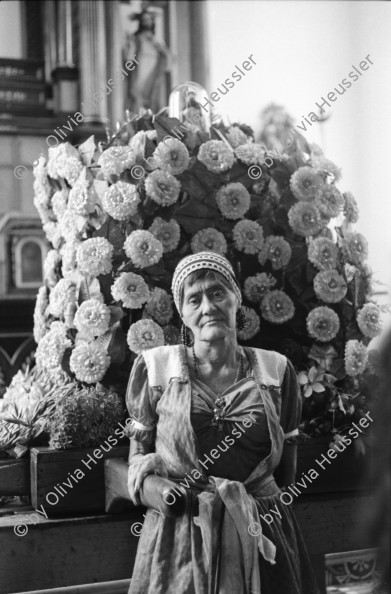 Image of sheet 19930520 photo 8: La vela de Santo Domingo, Iglesia de las Sierritas 9 de agosto 1993. Eine Schwester sitzt im Hintergrund.Sie überwacht aus Distanz die Feierlichkeiten die Tage später zu Toten führt , die in der Kirche ersticken. Der Dominguito ist ein kleiner Heiler und Heiliger unter einer Glashaube. Rausgeputzte Frau vor dem Blumenberg des Santos. Sie empfängt die Almosen und verteilt Blumen. Leute kommen auf den Knien zum Santos. Sie werden von einem mit Oel getränkten Mann geführt. Andenken und Glücksbringer werden verkauft. Managua Nicaragua 1993