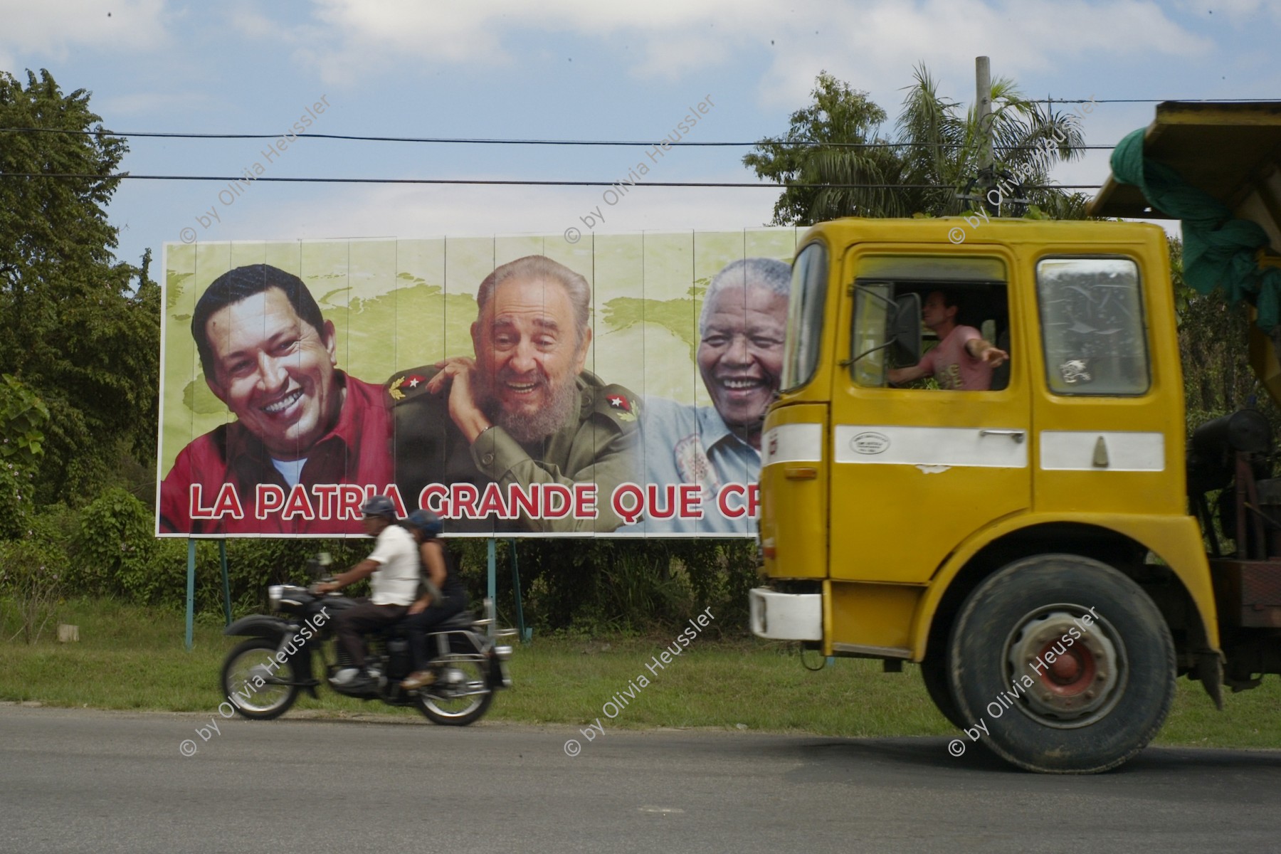 Image of sheet 20140214 photo 75: Lastwagen und Motorrad vor einem Plakat mit v.l.n.r. Hugo Chavez, Fidel Castro und Nelson Mandela, Cuba 2014.